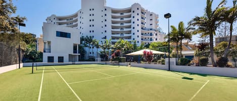 A tennis court with a net, surrounded by a fence, and a building with multiple balconies in the background.