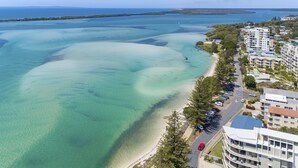 Eine Vogelperspektive einer Küstenlandschaft mit klarem türkisblauem Wasser, einem Sandstrand und einer Reihe mehrstöckiger Gebäude mit Balkonen.