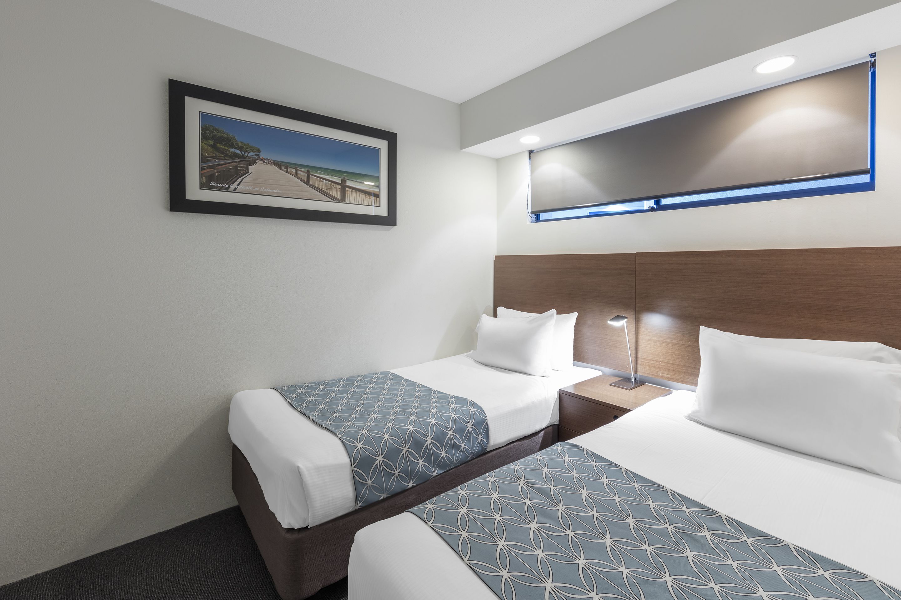 Et hotelværelse med to senge, et billede i ramme på væggen, et natbord med en lampe og et moderne loftsdesign.