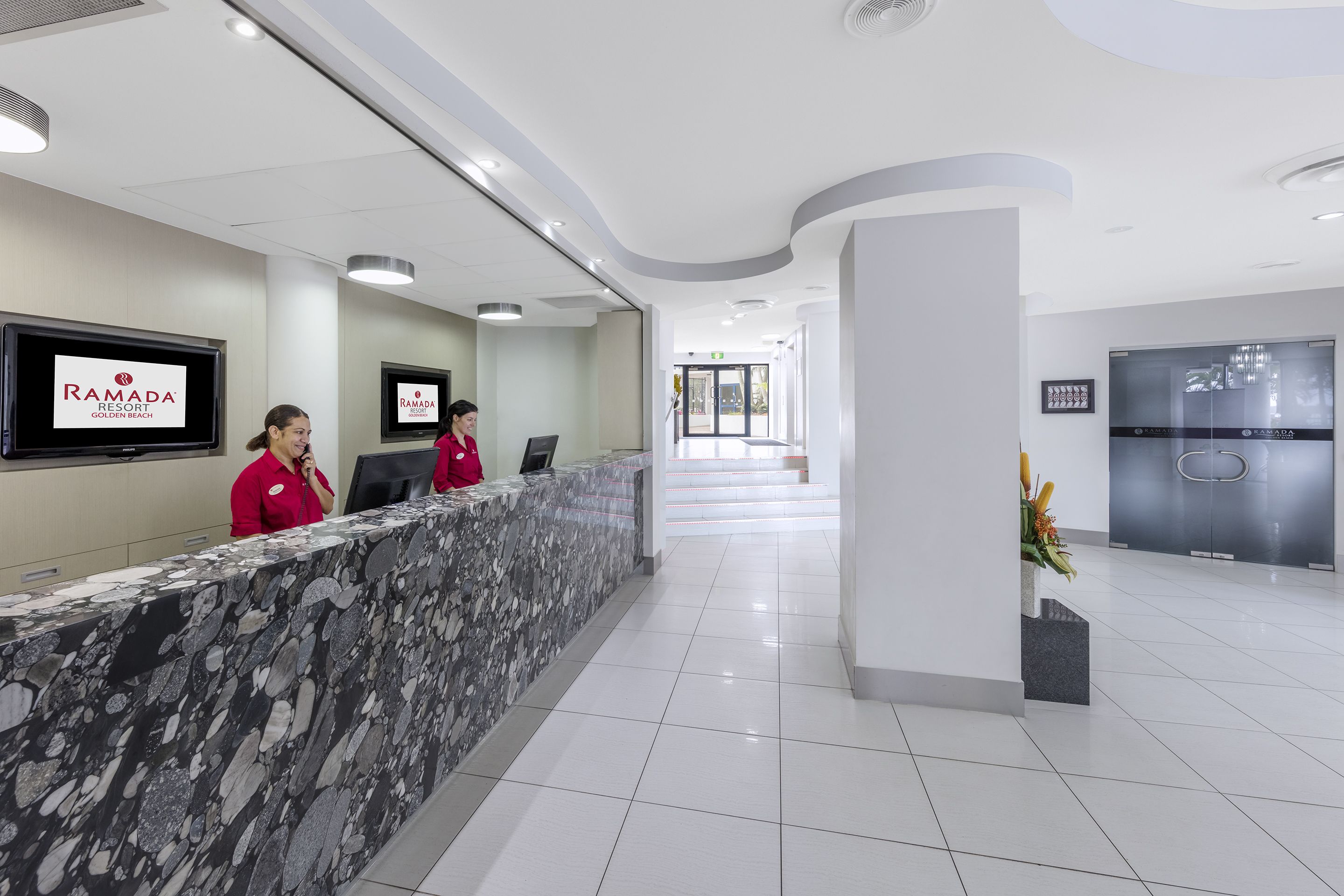 Zona de recepción con mostrador de mármol, dos recepcionistas con uniforme rojo, un televisor mostrando 'Ramada Golden Beach' y un diseño interior moderno.