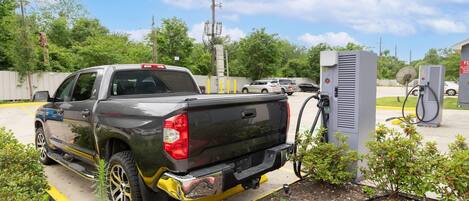 A pickup truck parked at a charging station in a parking lot with a fence, trees, and a clear sky in the background.