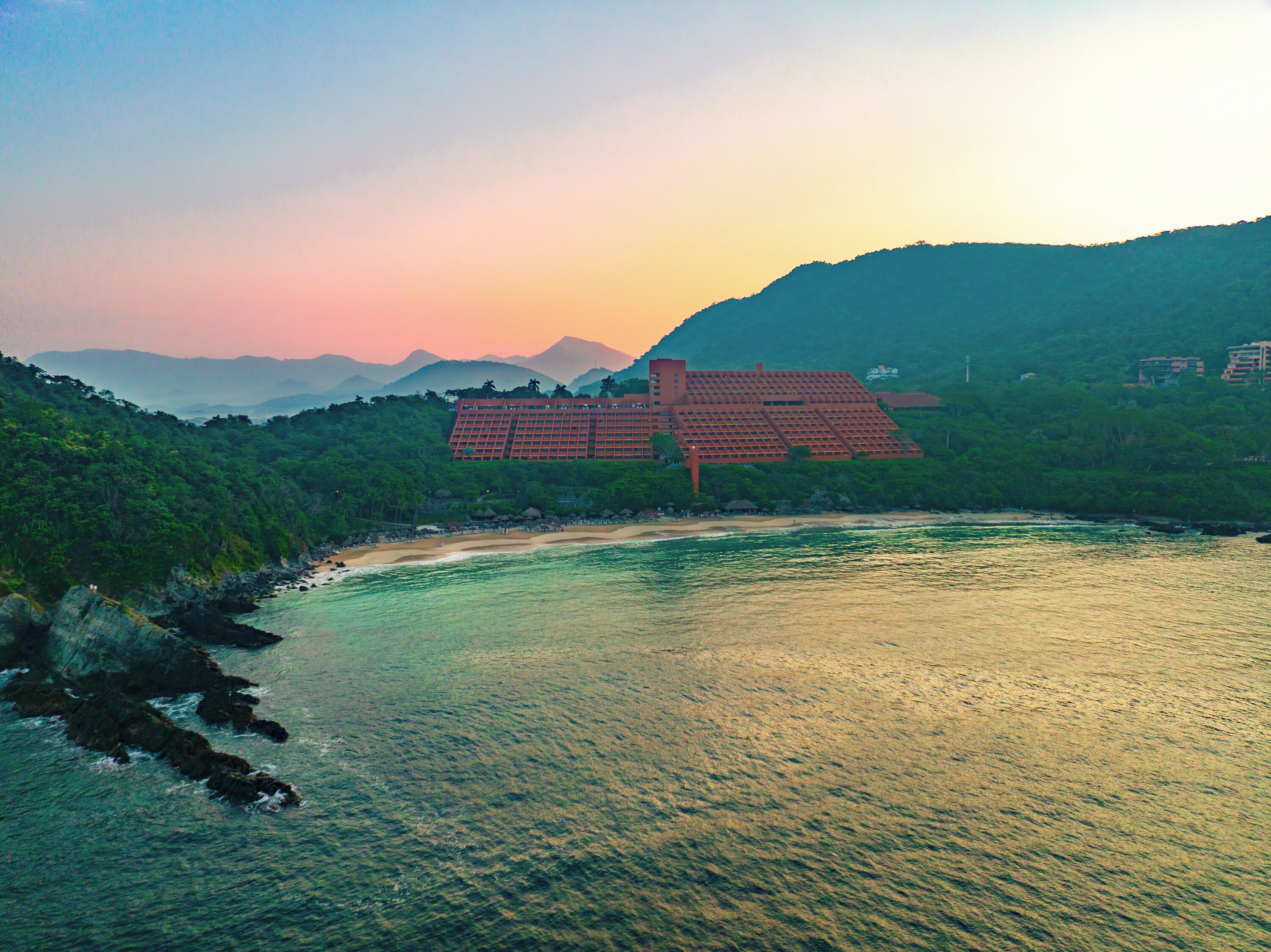 A large resort hotel with multiple buildings, situated by a beach and surrounded by lush greenery and mountains.