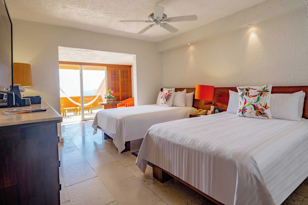 A hotel room with two beds, a desk with a lamp, a phone, and a window with a view of the outside.