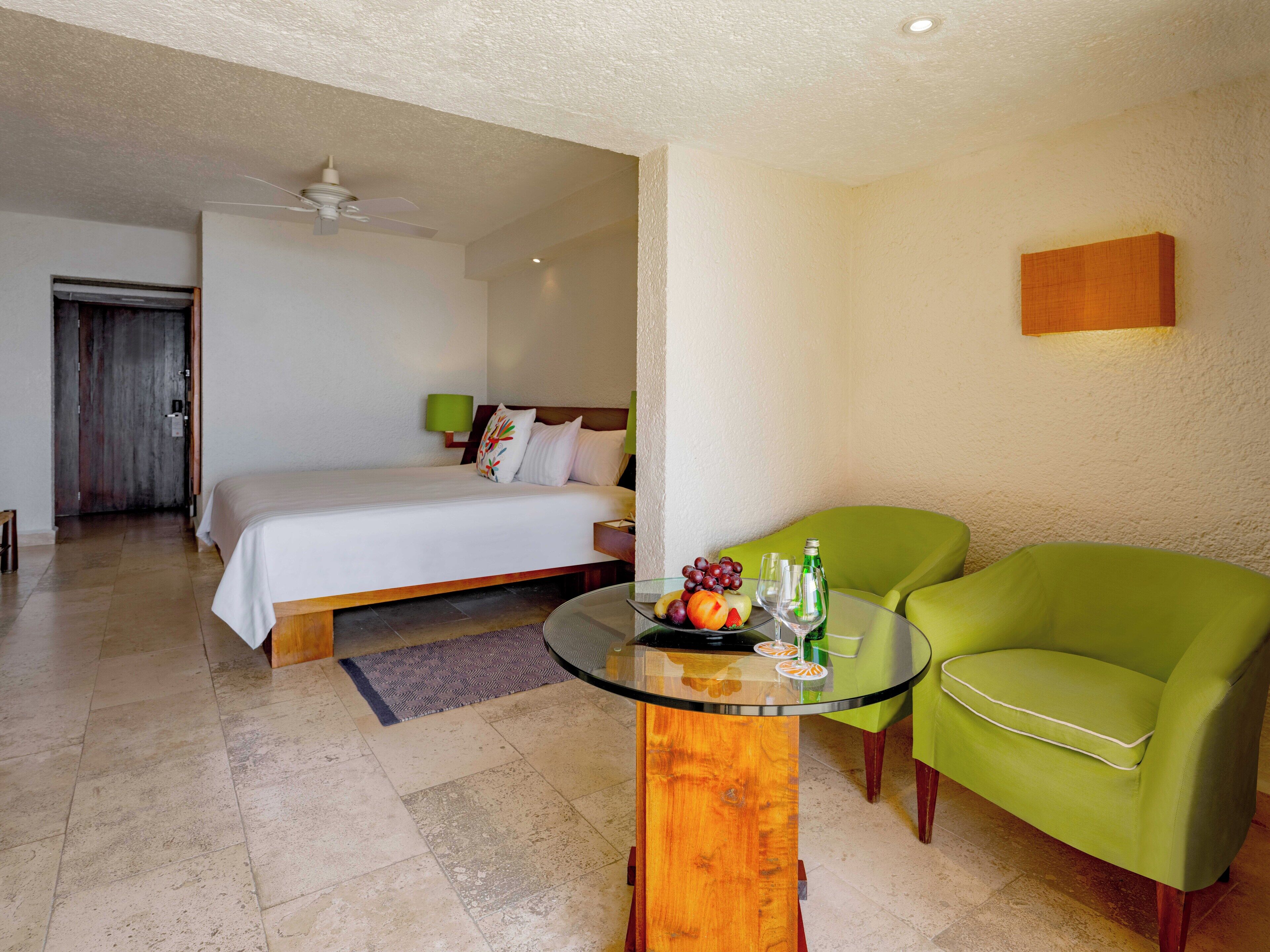 A hotel room with a bed, a green armchair, a glass coffee table, and a small table with fruit and glasses.