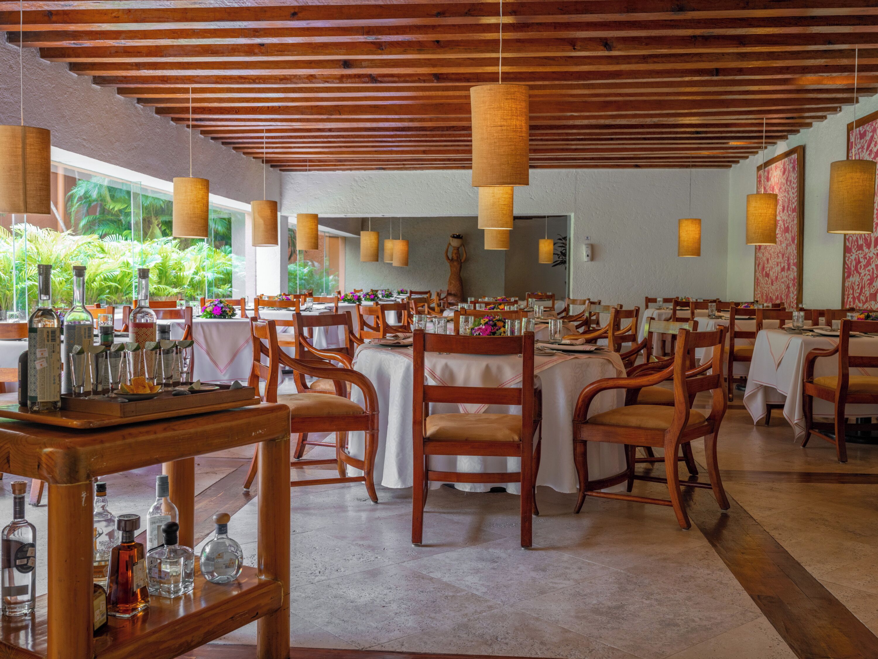 A restaurant with wooden tables and chairs, hanging pendant lights, and a view of greenery outside.