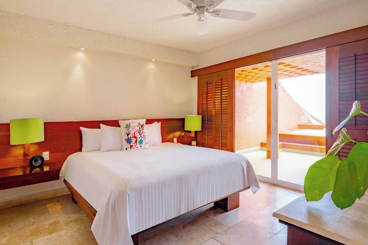 A hotel room with a bed, two bedside lamps, a ceiling fan, and a view of the outside balcony.