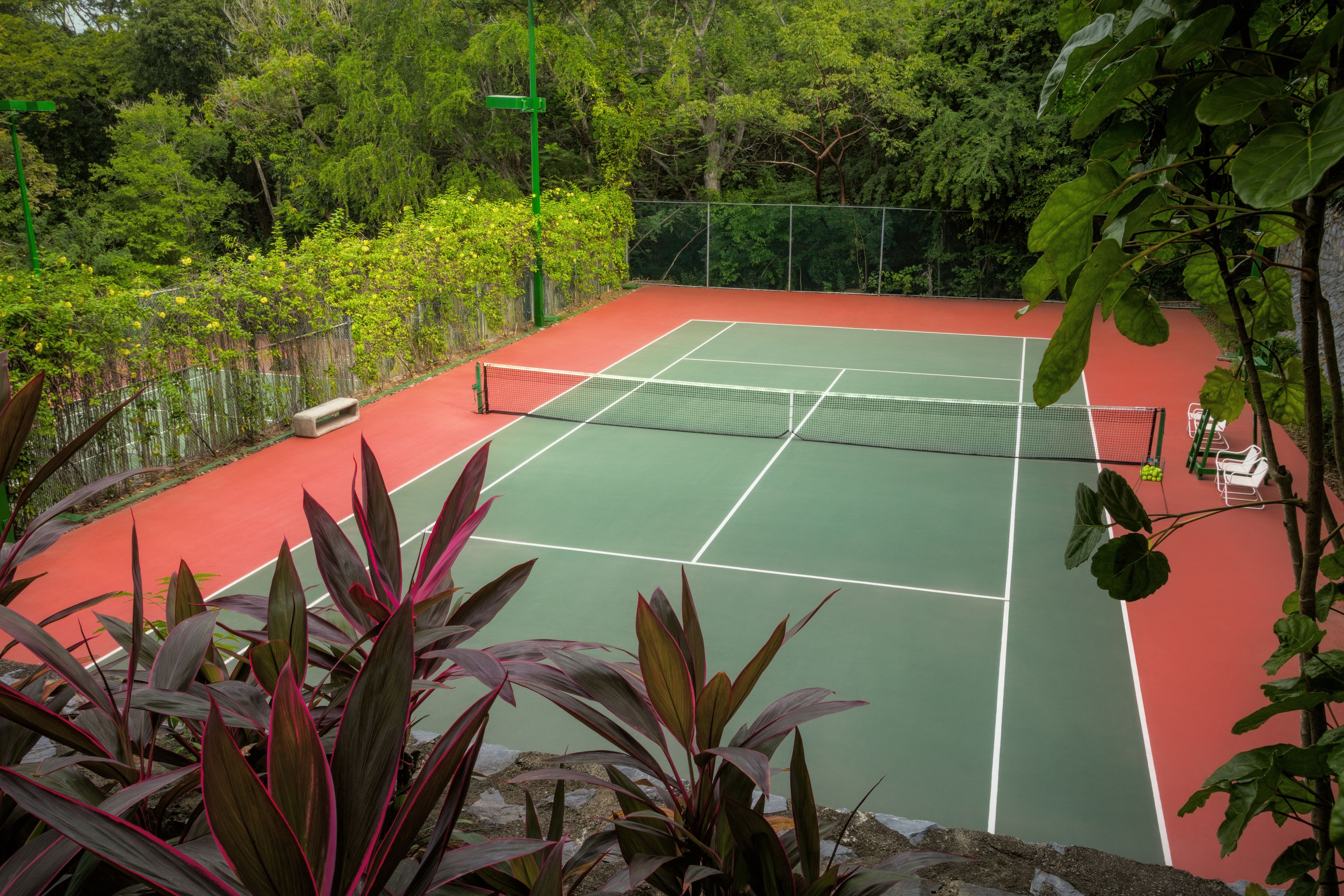 A tennis court surrounded by lush greenery and a bench.