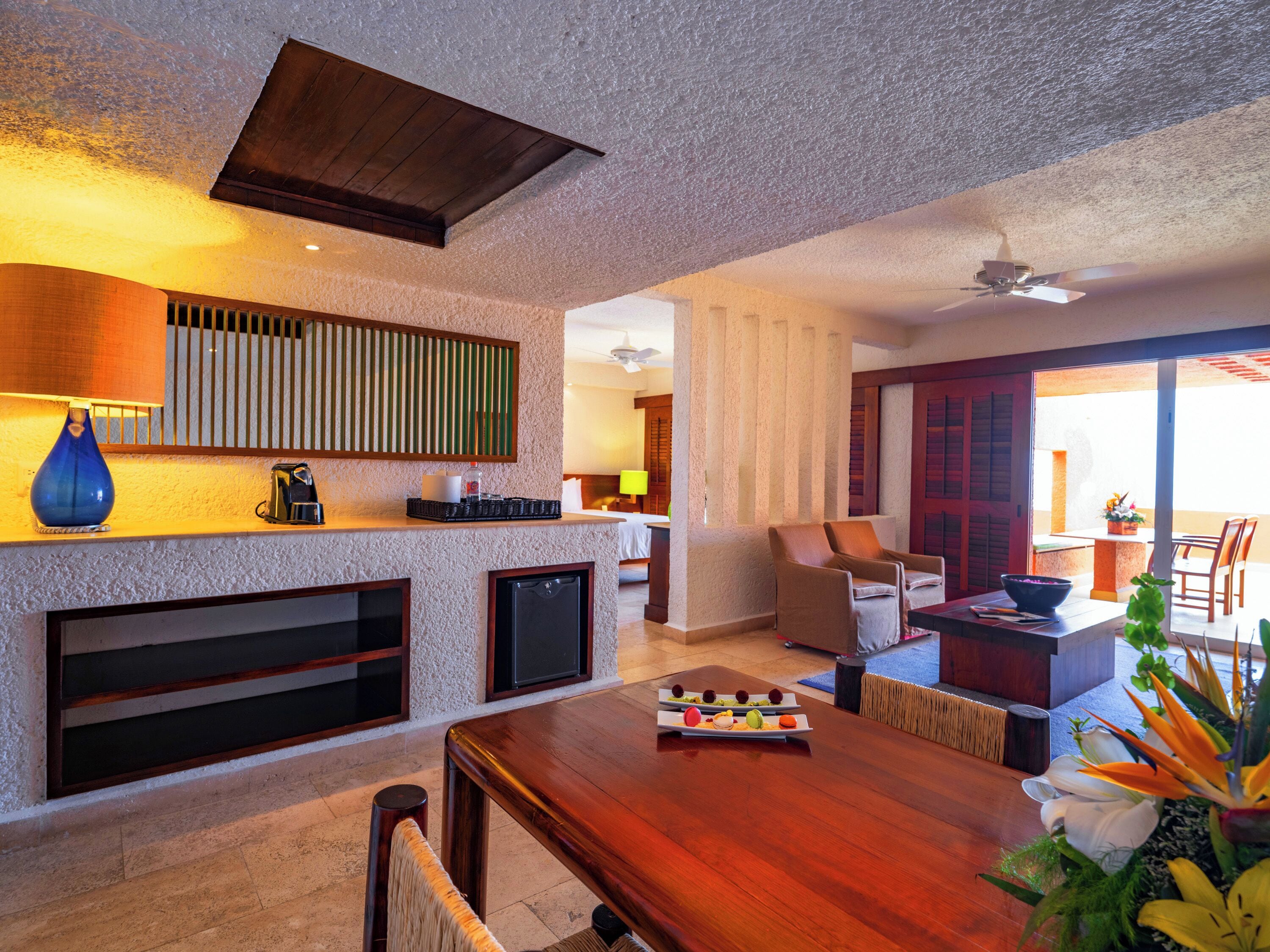 A modern hotel room with a wooden dining table, a kitchen area with a built-in refrigerator, and a large window offering a view of the outdoors.