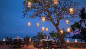An outdoor dining area with wicker hanging lights, a table set for two, and a view of the ocean at dusk.