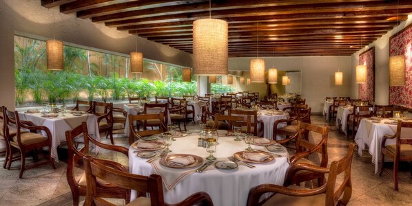 A restaurant with multiple round tables set for dining, wooden chairs, and hanging light fixtures.