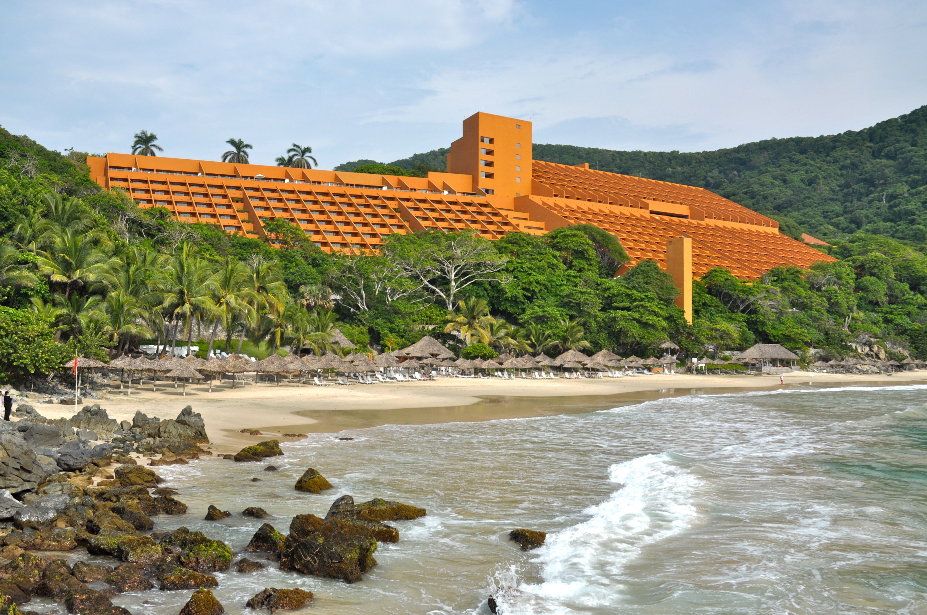 A beachfront hotel with a large building, palm trees, and a sandy beach.