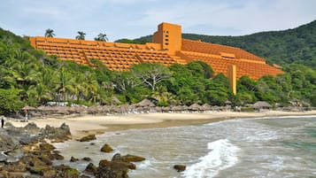 Un hotel frente a la playa con un edificio grande, palmeras y una playa de arena.