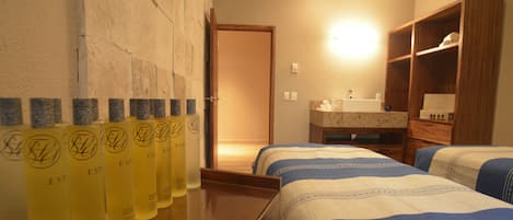 A hotel room with two beds, a wooden dresser, a bathroom with a sink and mirror, and a row of toiletries on the wall.