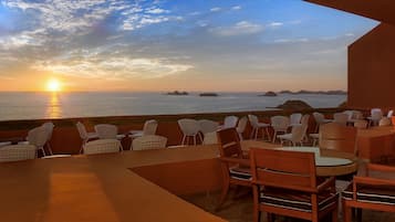 Et udendørs siddeområde med borde og stole med udsigt over havet i solnedgang.
