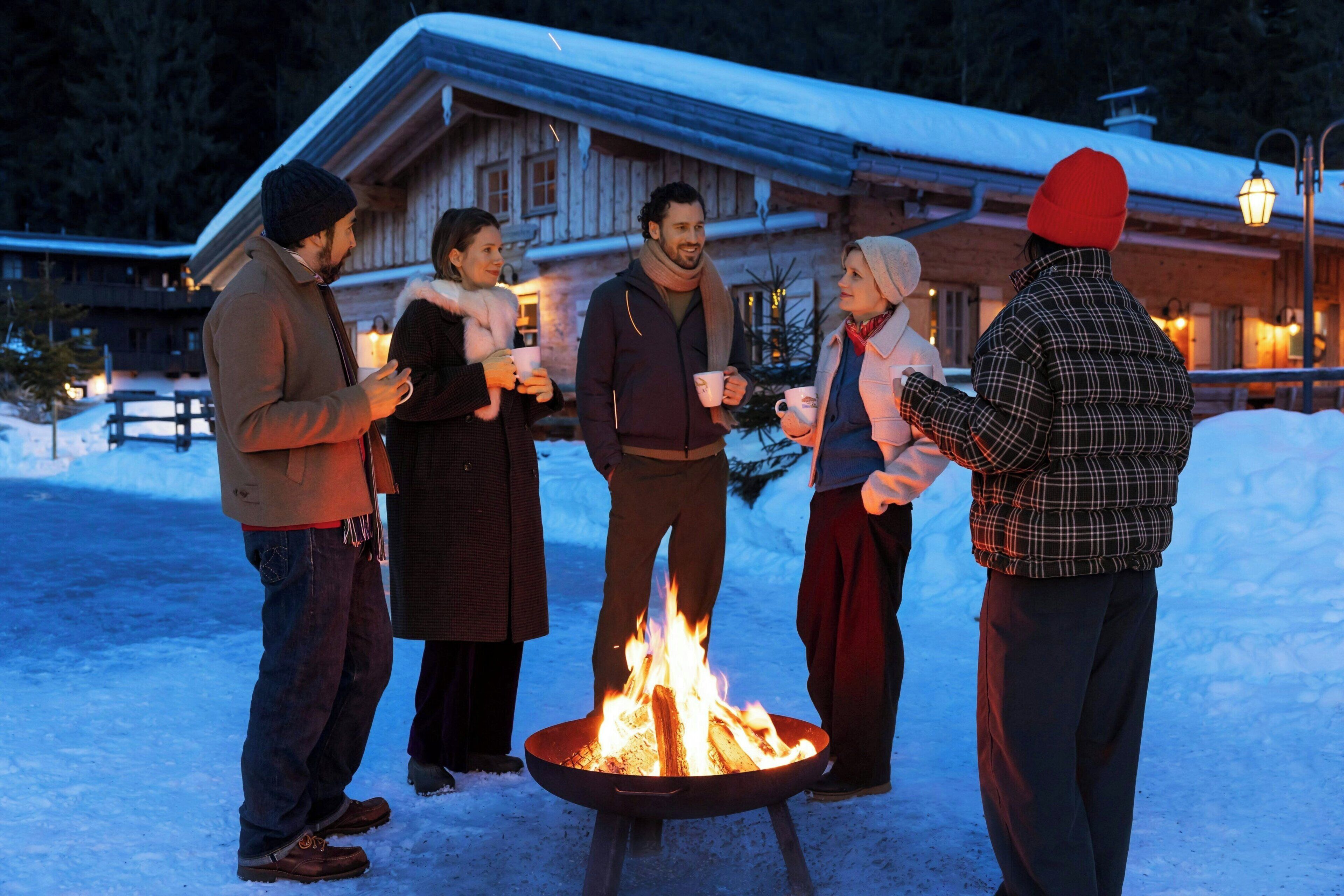 A group of people gathered around a fire pit in a snowy outdoor setting, with a wooden building in the background.
