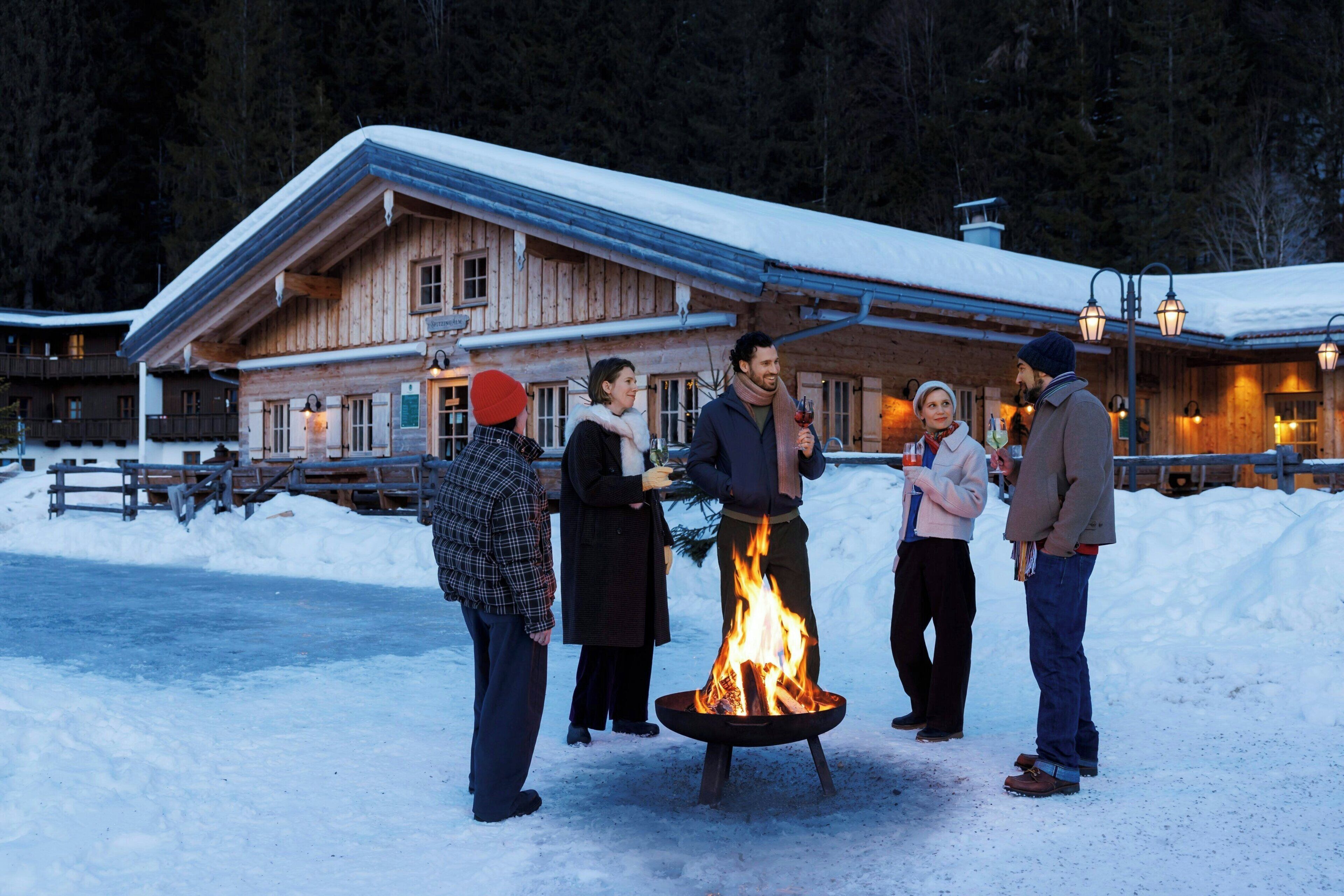 A wooden lodge with a fire pit in the foreground, surrounded by people.