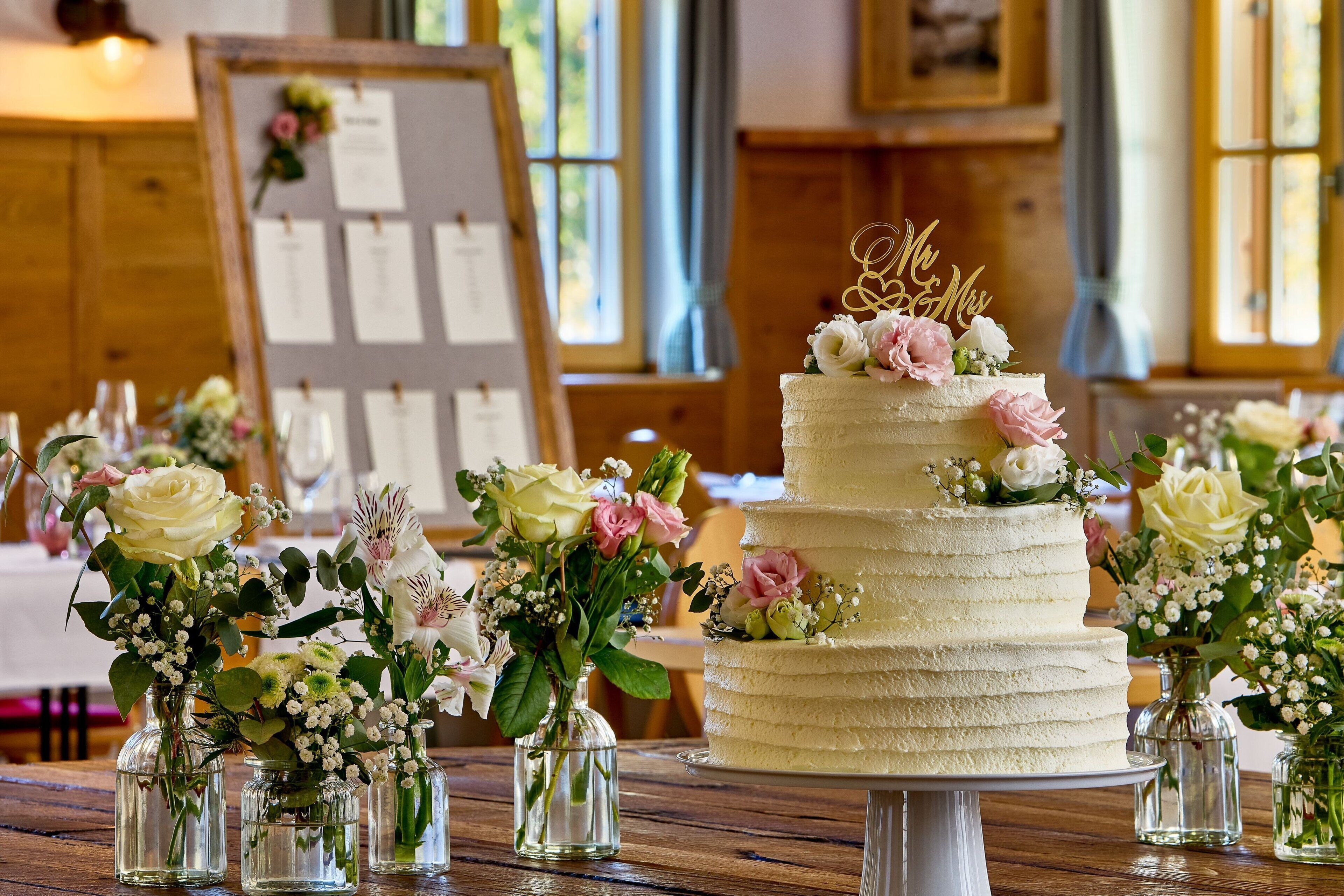 Ein mehrstöckiger Hochzeitstorte mit floraler Dekoration und einer Spitze, umgeben von kleinen Blumenvasen auf einem Holztisch.