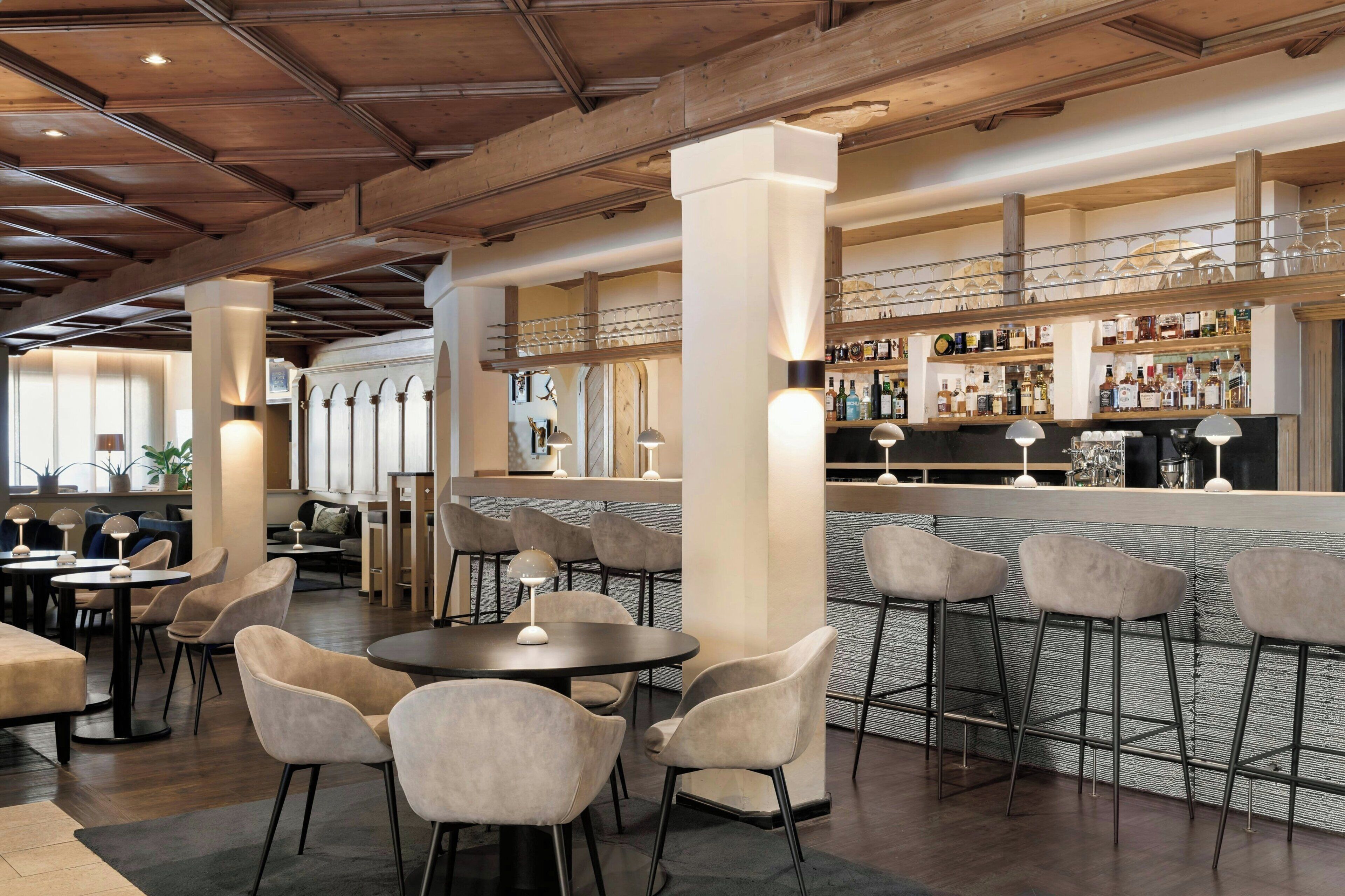 Un espace bar avec des tabourets hauts, un comptoir de bar orné d’étagères remplies de bouteilles, et un coin salon avec des tables et des chaises rondes.