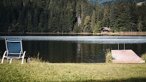 Vista de un lago con un muelle de madera, una silla y una cabaña al fondo.