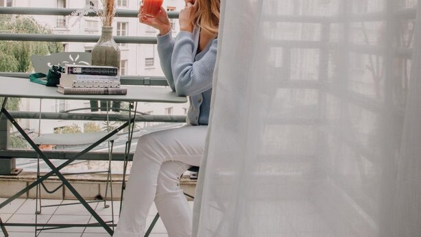 A person sitting on a balcony with a table, a laptop, and a drink. The balcony overlooks a row of buildings. - Pepper & Paper Apartments (Paris)