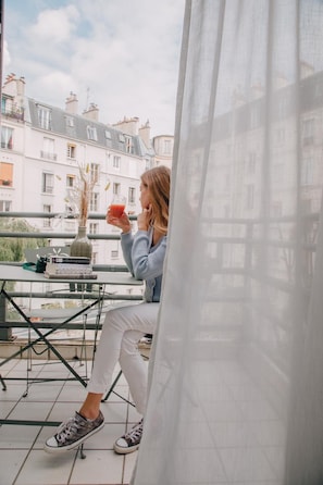 A person sitting on a balcony with a table, a laptop, and a drink. The balcony overlooks a row of buildings. - Pepper & Paper Apartments (Paris)
