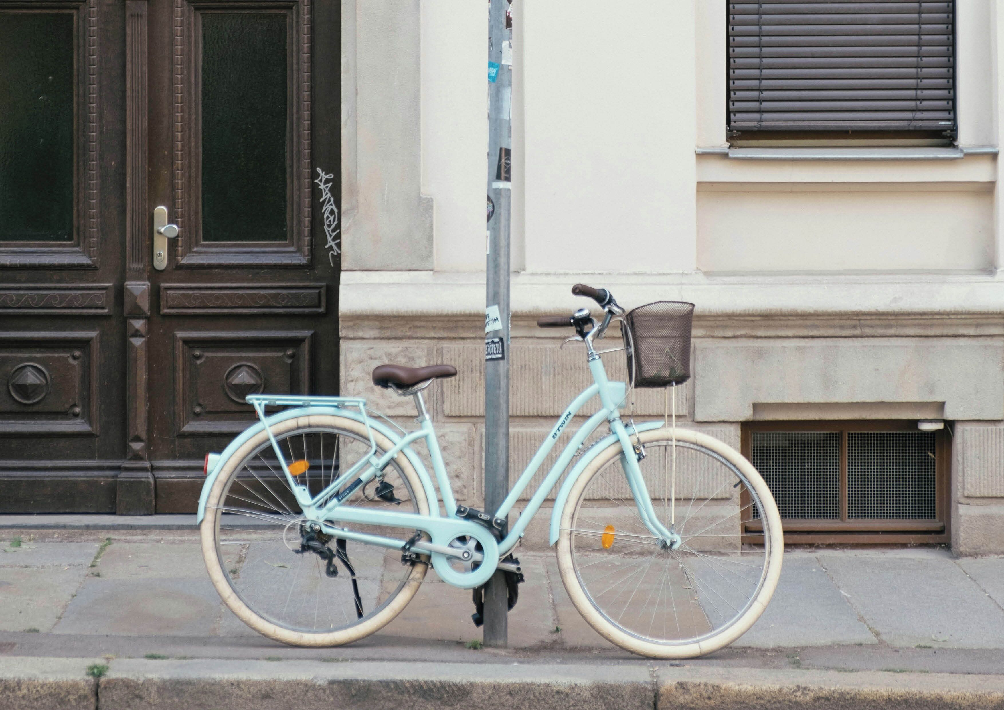 A light blue bicycle with a basket, parked on a sidewalk next to a door and a window.