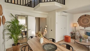 A modern living room with a wooden dining table, chairs, a sofa, and a coffee table. There is a staircase with a railing leading to an upper level. - Pepper & Paper Apartments (Paris)