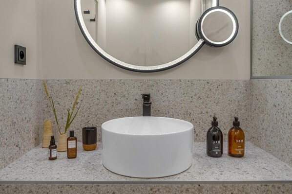 A bathroom with a round white sink, a mirror with a light, and various toiletries on the countertop. - Pepper & Paper Apartments (Paris)