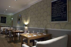 Un interior de restaurante con mesas y sillas de madera, un menú en una pizarra y un papel tapiz estampado.