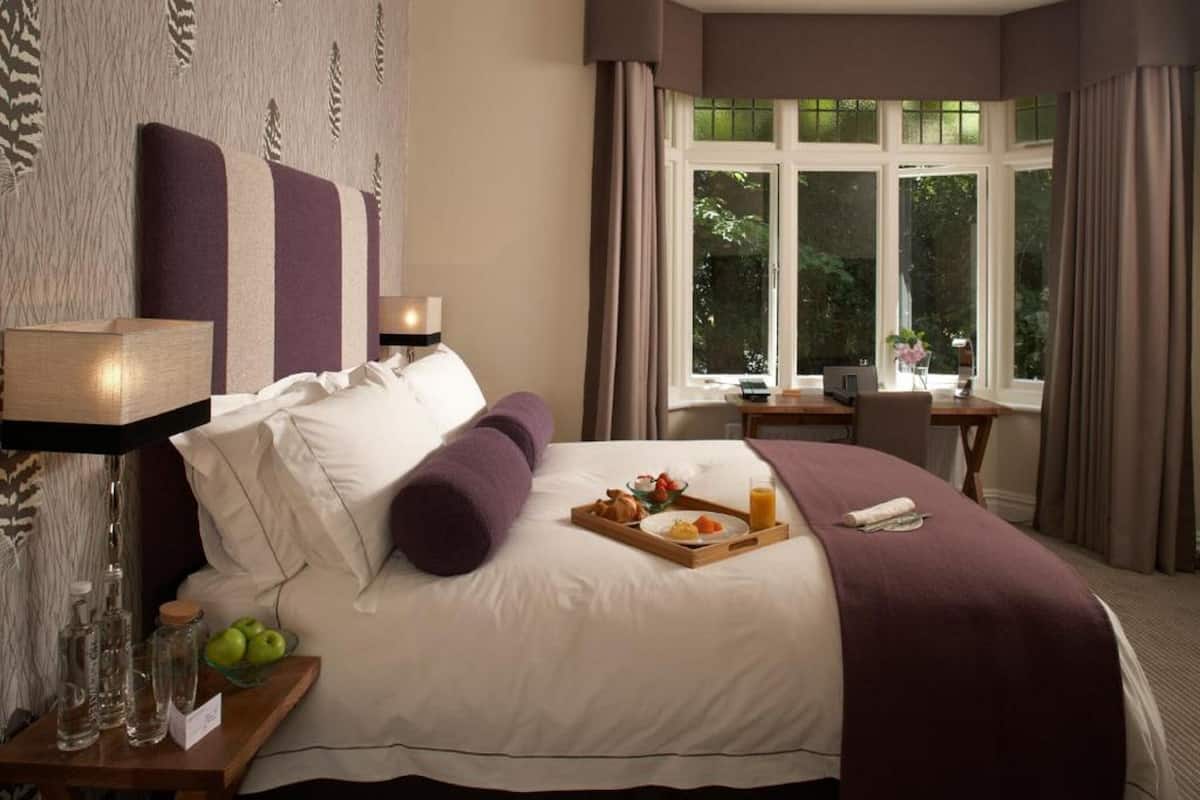 A hotel room with a large bed, a bedside table with a lamp, a tray with breakfast, and a window with curtains.