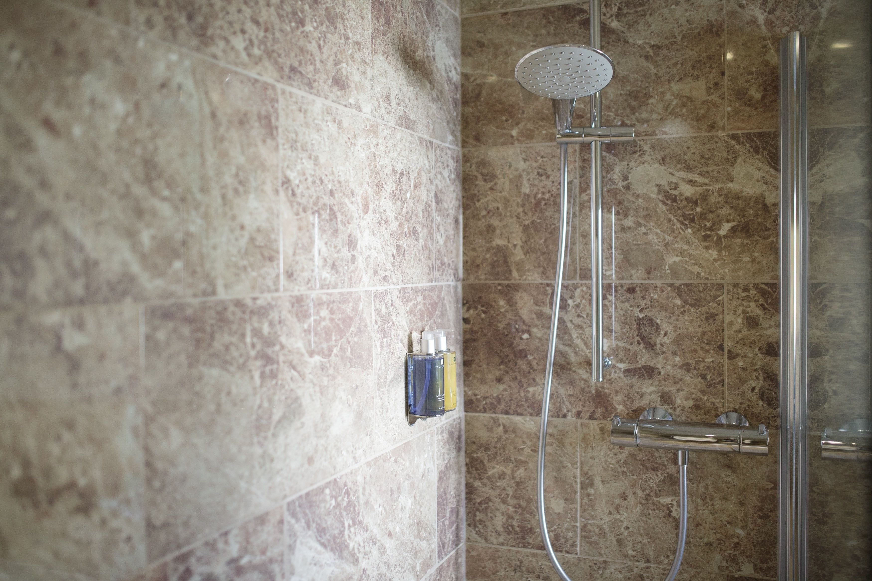 A shower with a handheld showerhead and a wall-mounted faucet, set against a marble wall.