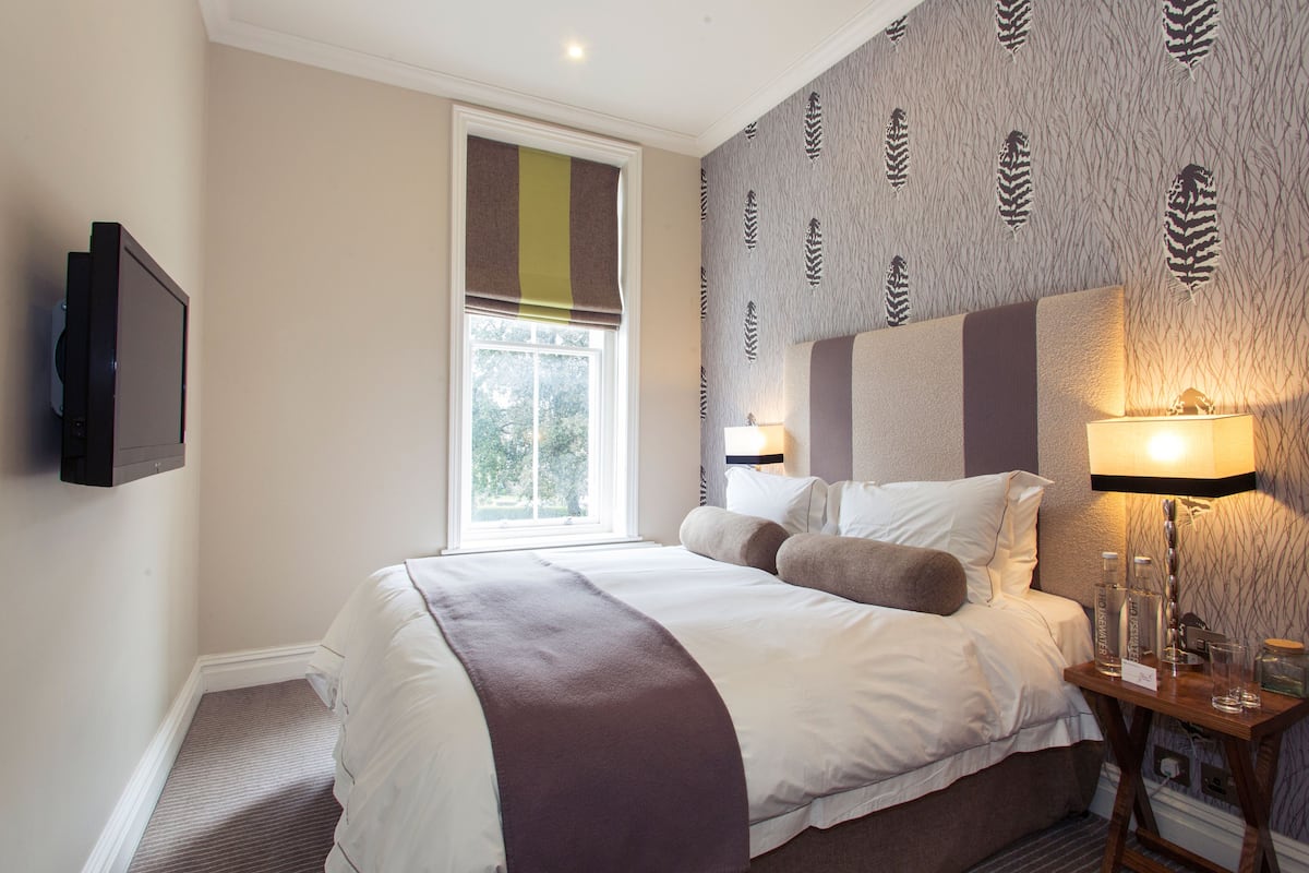 A hotel room with a large bed, a TV mounted on the wall, a window with a view of trees, and a nightstand with a lamp and a glass of water.