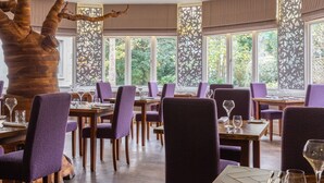 Un espace repas avec des tables en bois et des chaises à revêtement violet, de grandes fenêtres ornées de paravents décoratifs et une sculpture en forme d’arbre.