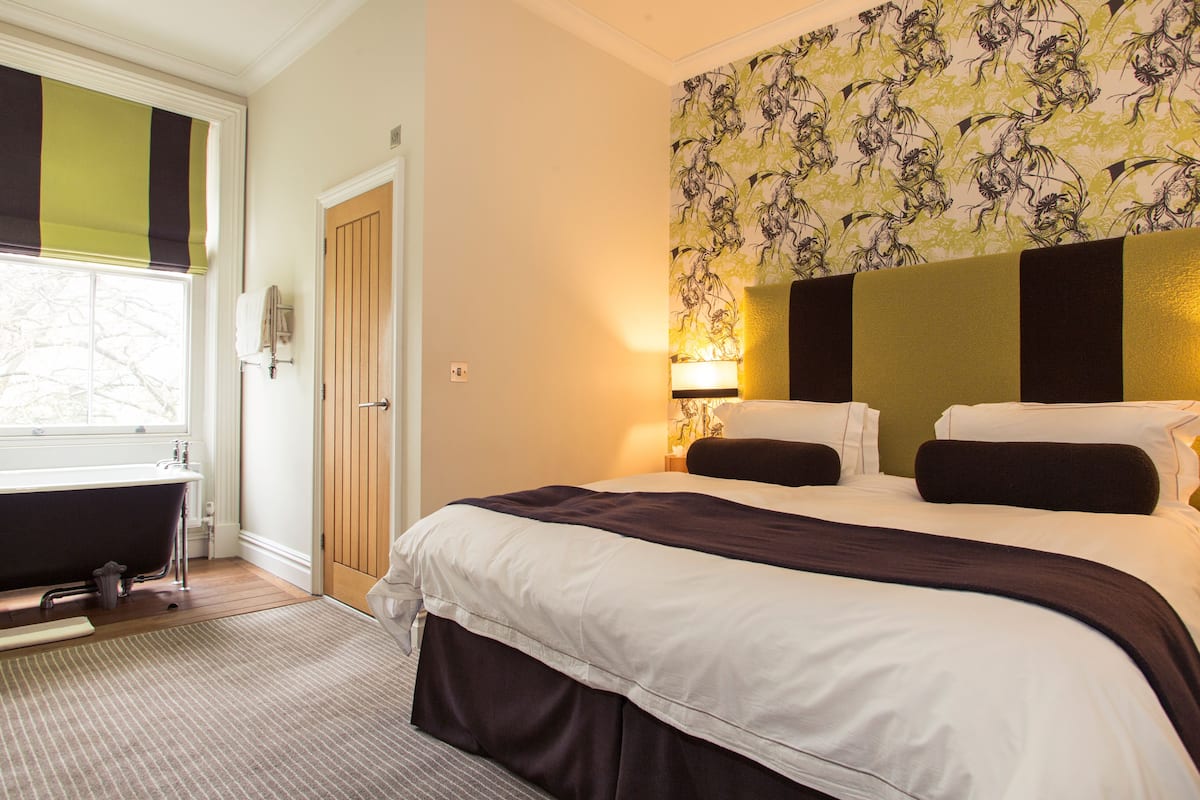 A hotel room with a large bed, a window with a view, a bathtub, and a patterned wallpaper.