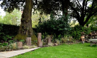 Un jardín con un banco de piedra, una escultura de madera y diversas plantas.