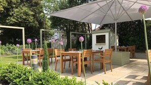 Un espace extérieur aménagé en terrasse, avec des tables et des chaises en bois, un grand parasol et une petite structure dotée d’une fenêtre.