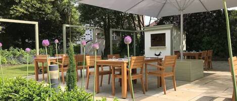An outdoor patio area with wooden tables and chairs, a large umbrella, and a small structure with a window.