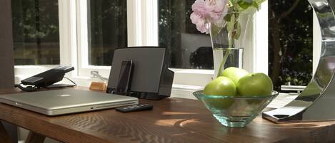 A wooden desk with a laptop, a phone, a remote, a bowl of green apples, and a vase with flowers.