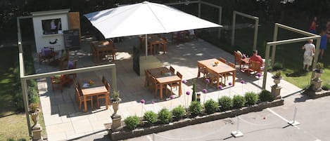An outdoor patio area with wooden tables and chairs, a white umbrella, and a small kiosk.