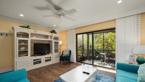 Ein Wohnzimmer mit Fernseher, weißem Unterhaltungsschrank, blauen Sofas und einer großen Glasschiebetür zu einer Terrassenanlage im Freien.