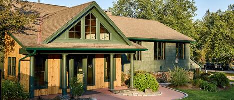 A two-story house with a green roof and wooden siding, featuring a covered front porch and a landscaped front yard.