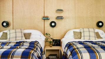 Quarto com duas camas, cada uma com um cobertor xadrez, uma mesinha de cabeceira com livros, um vaso com uma planta e um relógio fixado na parede.