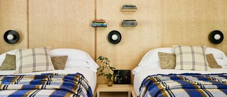 Una habitación con dos camas, cada una con una manta escocesa, una mesita de noche con libros, un jarrón con una planta y un reloj en la pared.