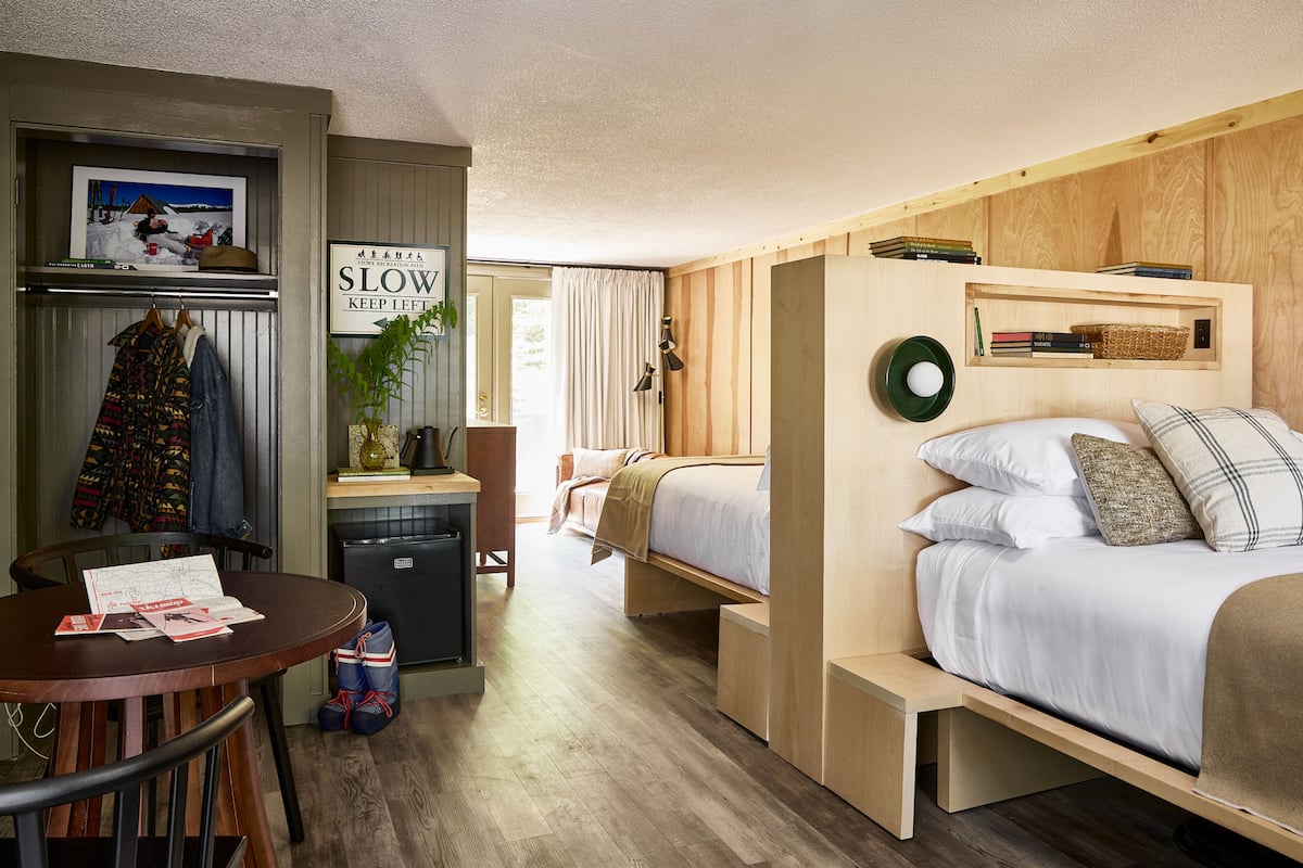 Pieni hotellihuone, jossa on sänky, pieni työpöytä, tuoli, pöytä ja pieni keittiönurkkaus.