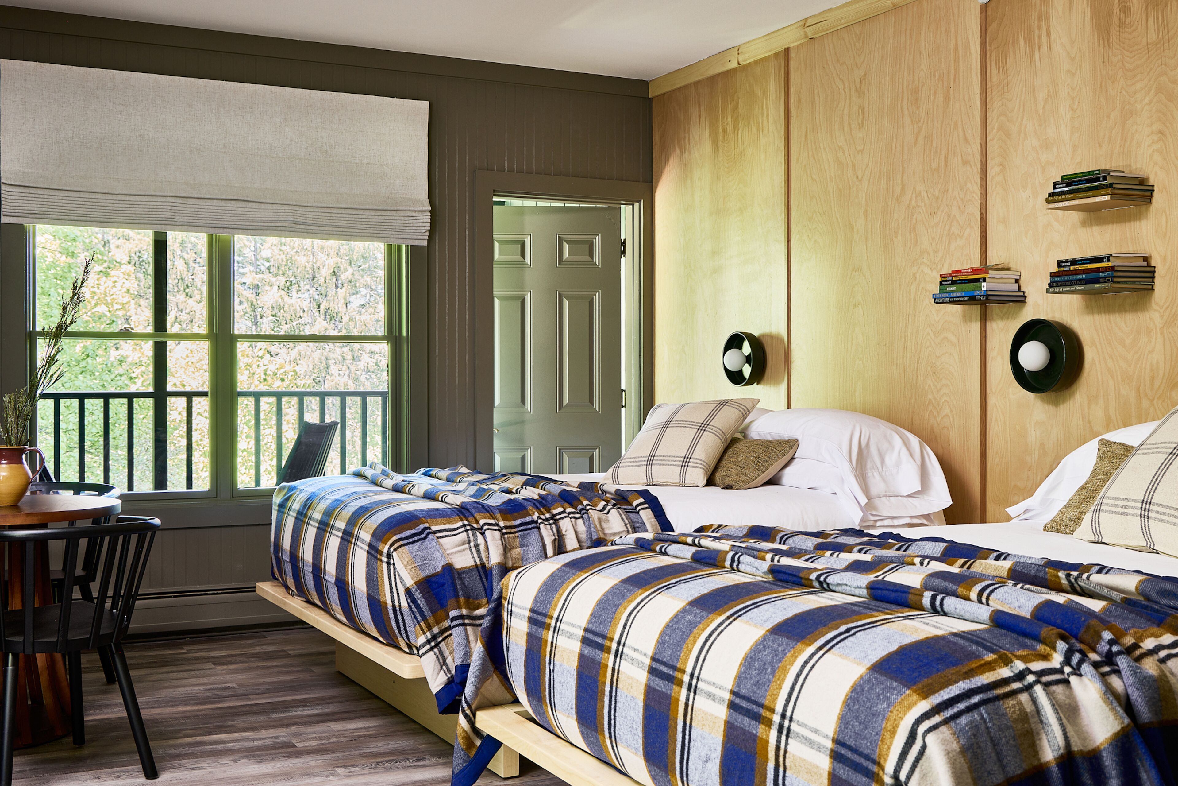 A bedroom with two beds, a wooden wardrobe, a window with blinds, and a small table with a plant.