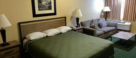 A hotel room with a bed, a sofa, a desk, a lamp, a picture on the wall, and an air conditioning unit.