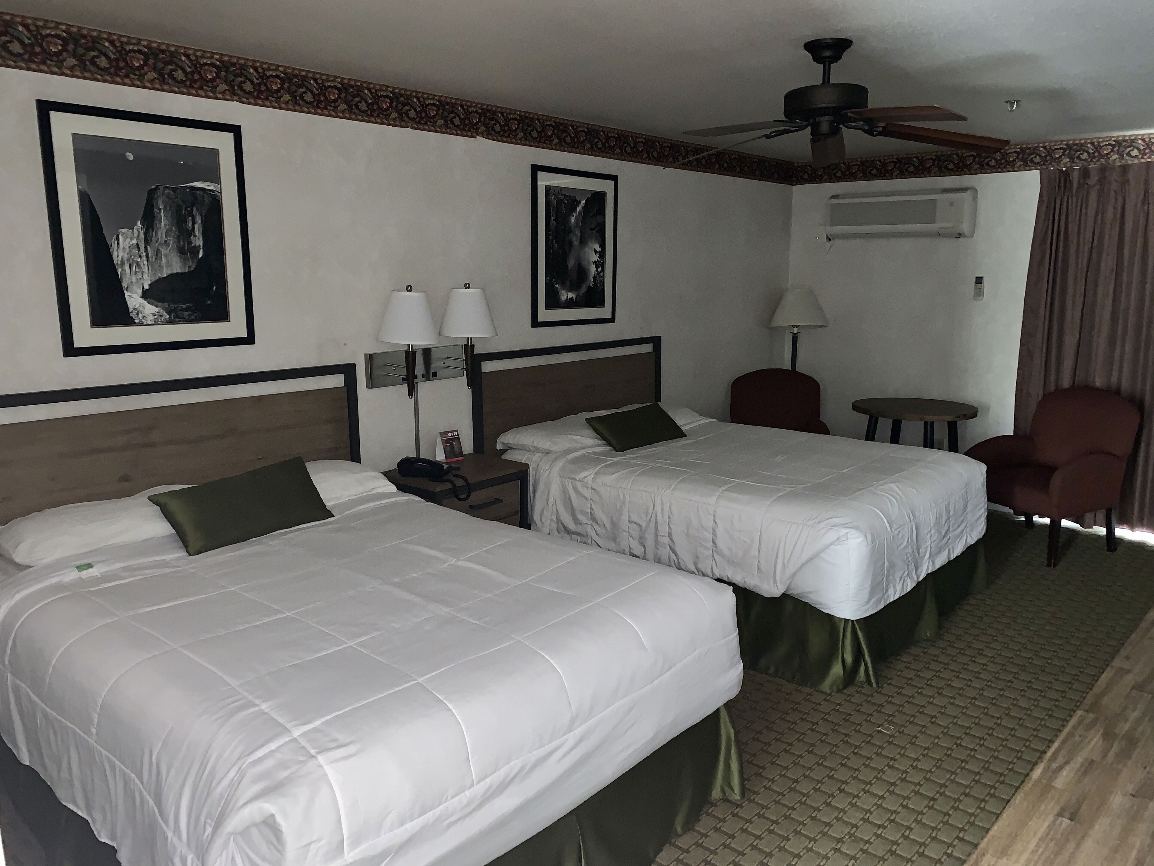 A hotel room with two single beds, a desk with a chair, a small table, a ceiling fan, and framed pictures on the wall.
