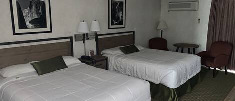 A hotel room with two single beds, a desk with a chair, a small table, a ceiling fan, and framed pictures on the wall.