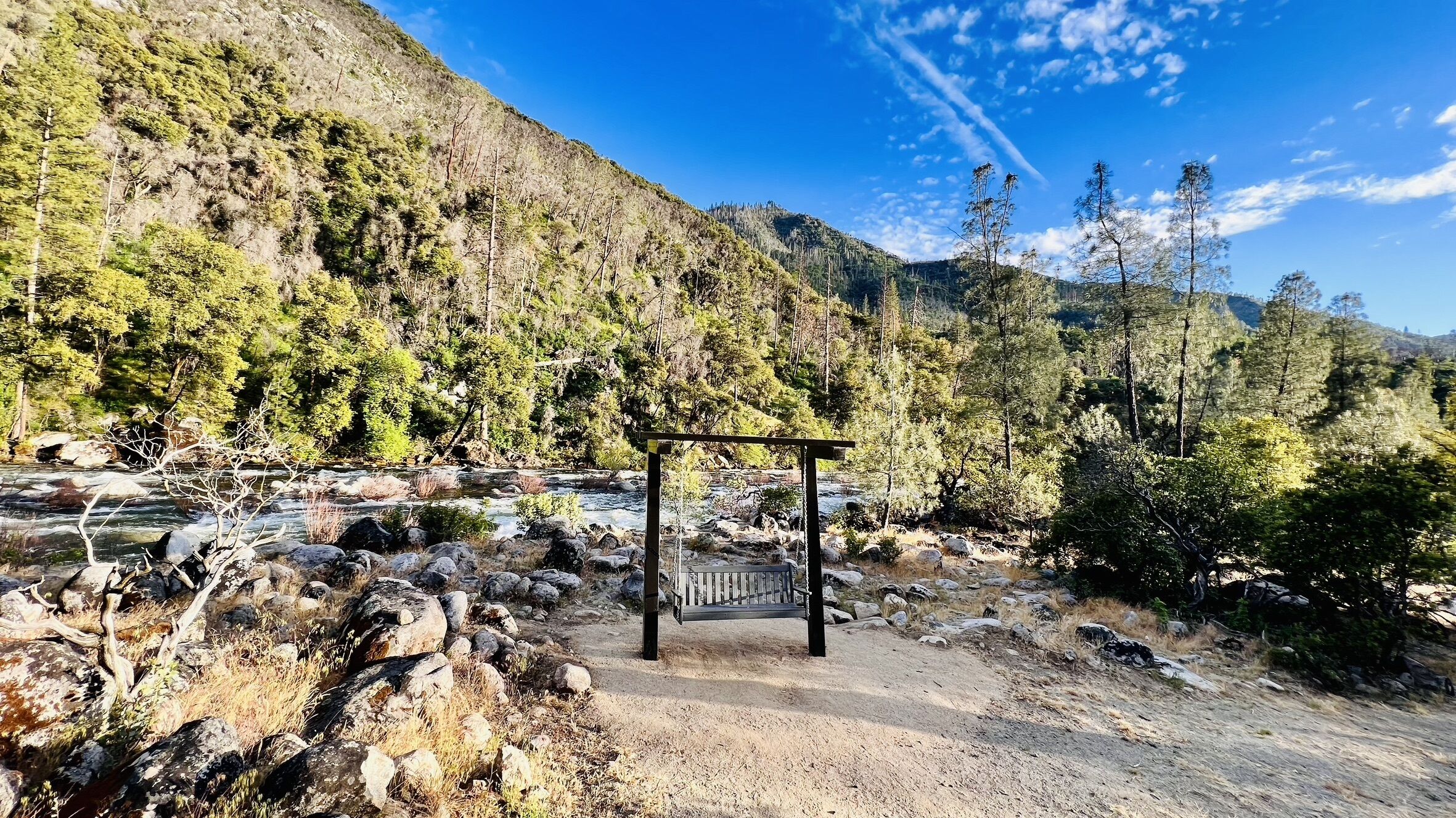 A wooden bench in a natural setting with a river and forested hills in the background.