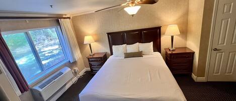 A hotel room with a large bed, two bedside lamps, a ceiling fan, a window with curtains, and a door.